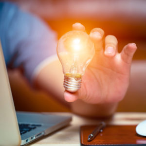 A person's hand holding a light bulb, which symbolically represents an idea, and with a laptop beside him, showing that he is working on that idea. This summarizes how to undertake.